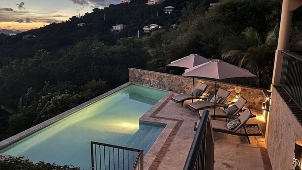 Una piscina amb gandules i ombrelles, envoltada per una paret de pedra i amb vistes a un turó on hi ha cases.