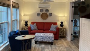 A cozy living room with a red sofa, blue armchair, patterned ottoman, and a ceiling fan. - Beach Walk Bungalow 1Bedroom 1Bath, 4 blocks to the beach!! Private Courtyard (St. Simons Island)