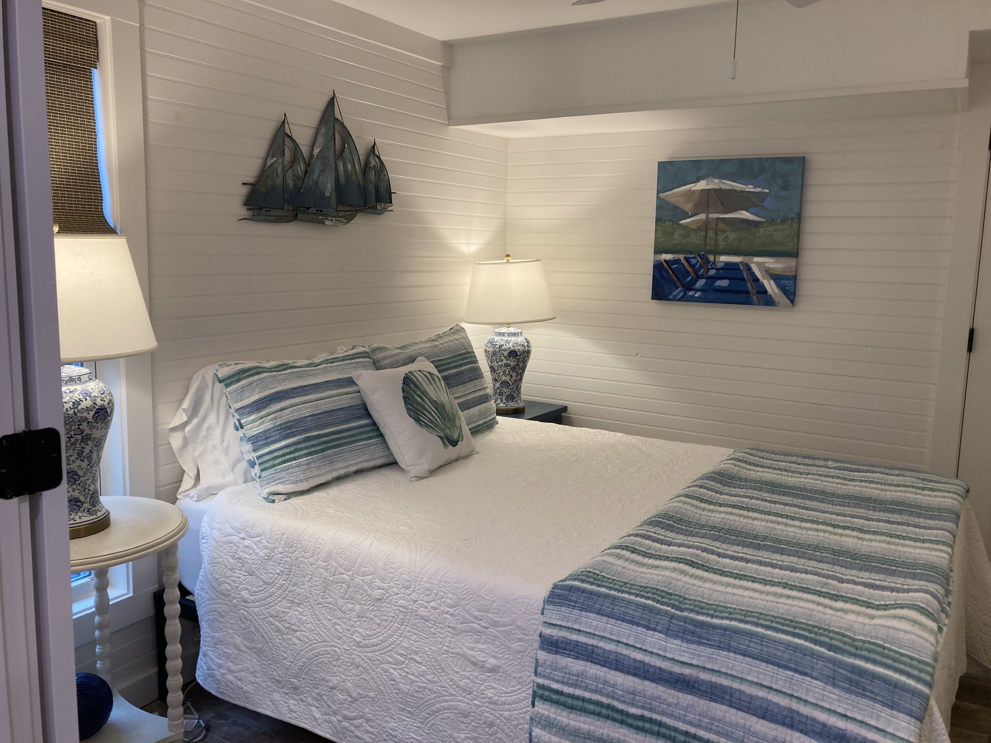 A neatly made bed with striped bedding, two bedside lamps, a decorative wall piece, and a painting of a boat.