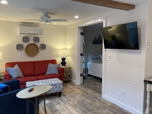 Un salon chaleureux avec un canapé rouge, des coussins à motifs, une petite table ronde et un ventilateur de plafond. Un téléviseur est fixé au mur, et on aperçoit un lit dans la pièce adjacente.