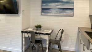 A kitchen with white paneling, a painting of the ocean, a TV, and a dining table with chairs. - Beach Walk Bungalow 1Bedroom 1Bath, 4 blocks to the beach!! Private Courtyard (St. Simons Island)