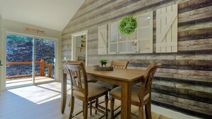 Uma área de refeições com mesa e cadeiras de madeira, vista através de uma porta de vidro para um deck externo, e uma parede rústica com detalhes decorativos.