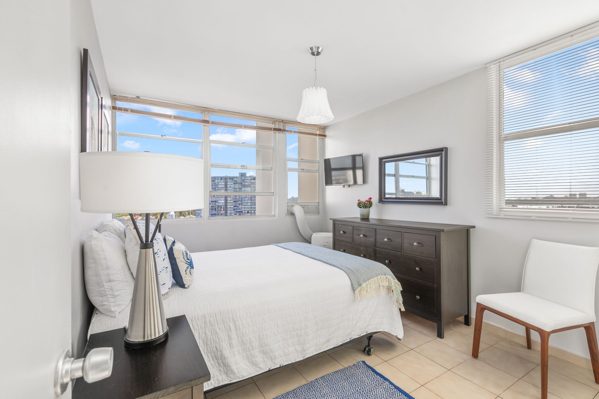 A bedroom with a bed, a dresser, a chair, a TV, and a window with blinds.