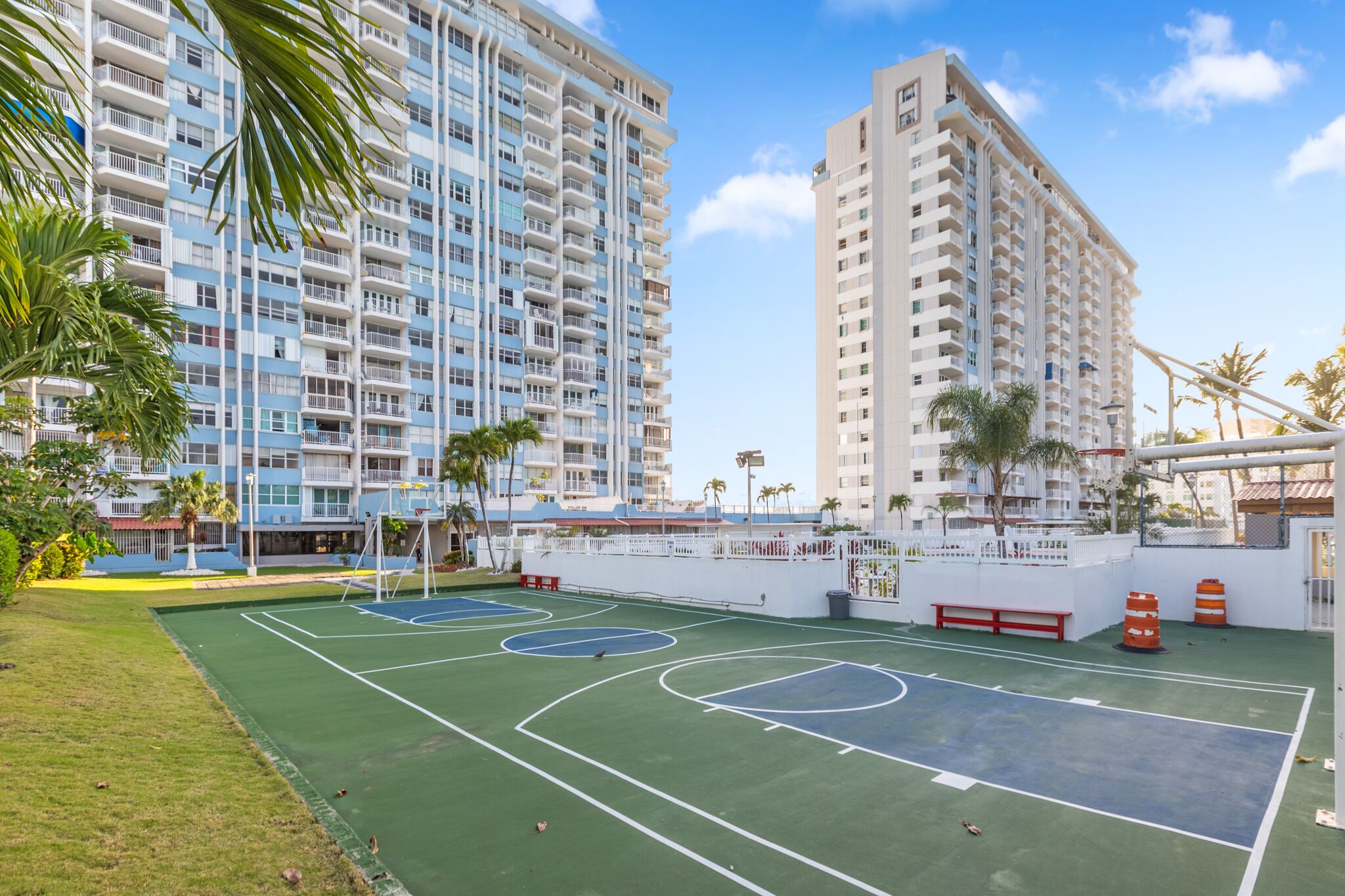 A basketball court surrounded by high-rise buildings, palm trees, and a white fence.