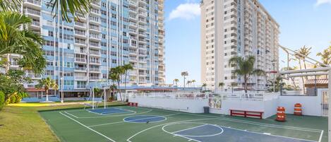 A basketball court surrounded by high-rise buildings, palm trees, and a white fence.