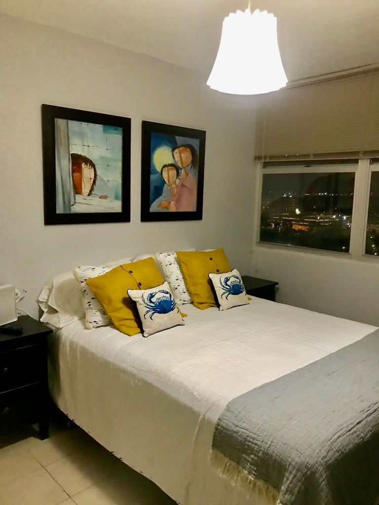 A bedroom with a bed, two paintings, a nightstand, a lamp, and a window with a city view.