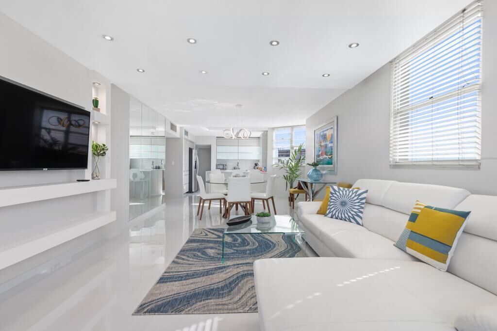 A modern living room with a flat-screen TV, a white sofa with decorative pillows, a glass coffee table, and a patterned rug.