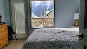 Un dormitorio con cama, cómoda, silla, ventana con vistas a montañas y puerta.