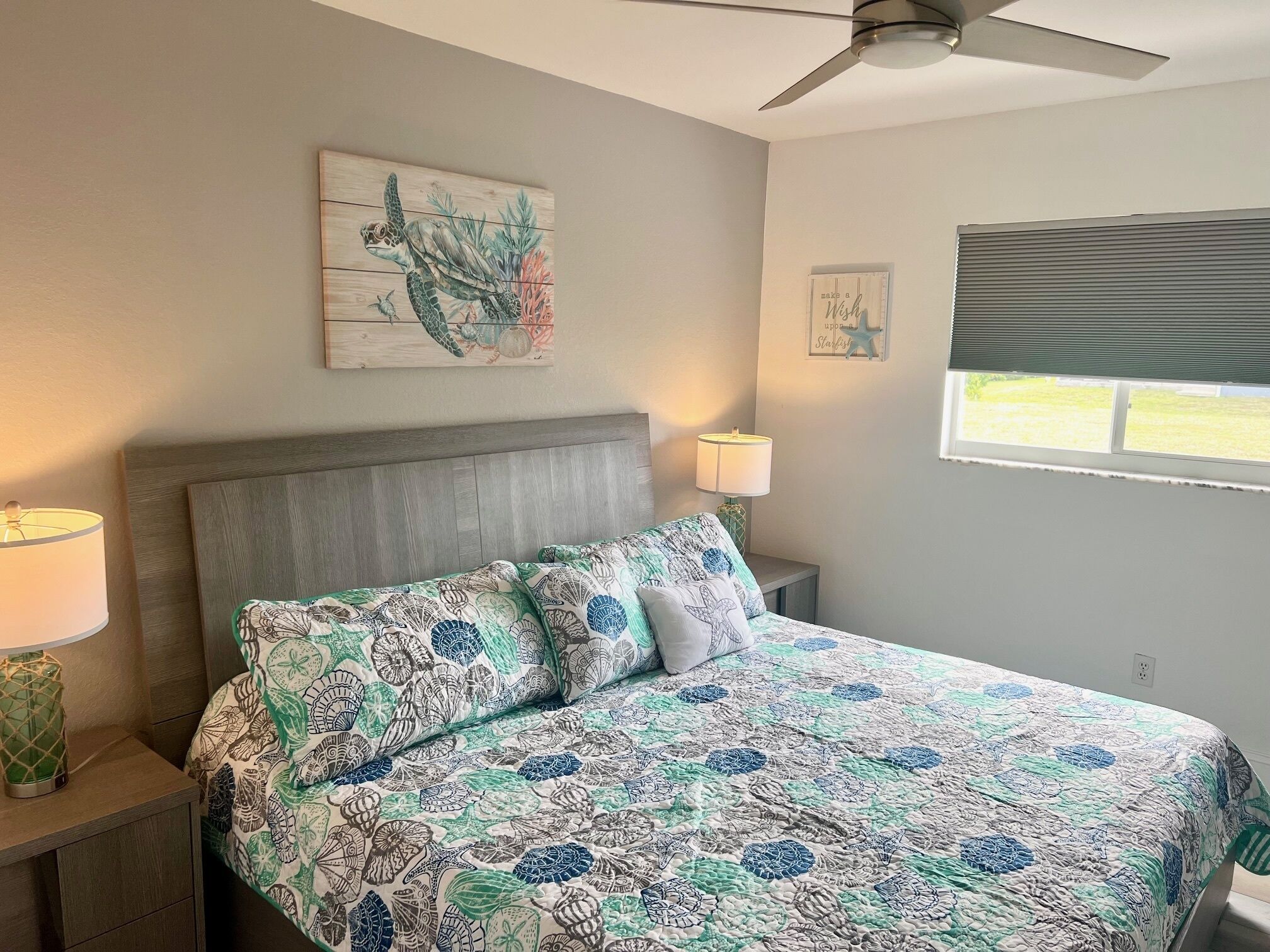A bedroom with a bed, two bedside lamps, a ceiling fan, a window with blinds, and a wall art piece.
