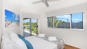 Ein Schlafzimmer mit Bett, Deckenventilator, großem Fenster mit Blick ins Freie und einer Tür, die auf einen Balkon führt.