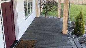 Una terraza de madera con piso oscuro, una puerta roja y un pequeño árbol al fondo.