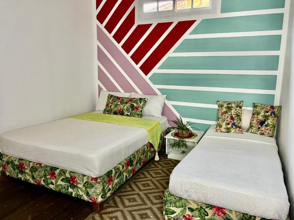 A room with two beds, one with a floral bedspread and the other with a plain white bedspread. There is a small table with a plant and a decorative item on it. The walls have a geometric pattern with red and white stripes.