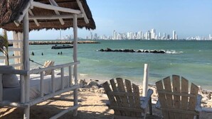 Una platja amb una cabana de palla, dues cadires de fusta i un paisatge urbà al fons.
