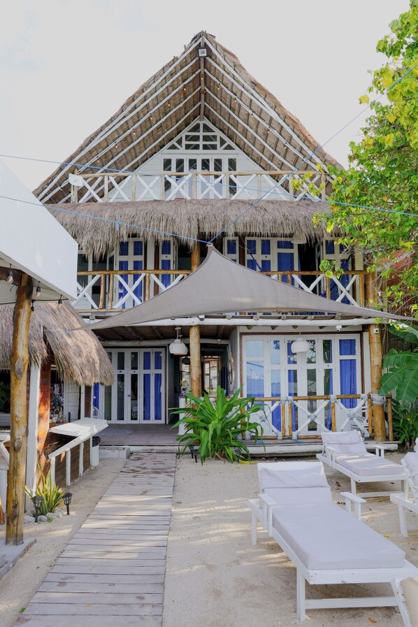 Un hôtel en bord de mer, doté d’un toit de chaume, de murs blancs et de touches de couleur bleue. Il dispose d’un espace extérieur couvert avec des fauteuils et d’une terrasse en bois menant à l’entrée.
