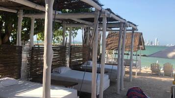 Eine Strandhütte mit strohgedecktem Dach, weißen Liegestühlen und sandigem Untergrund.