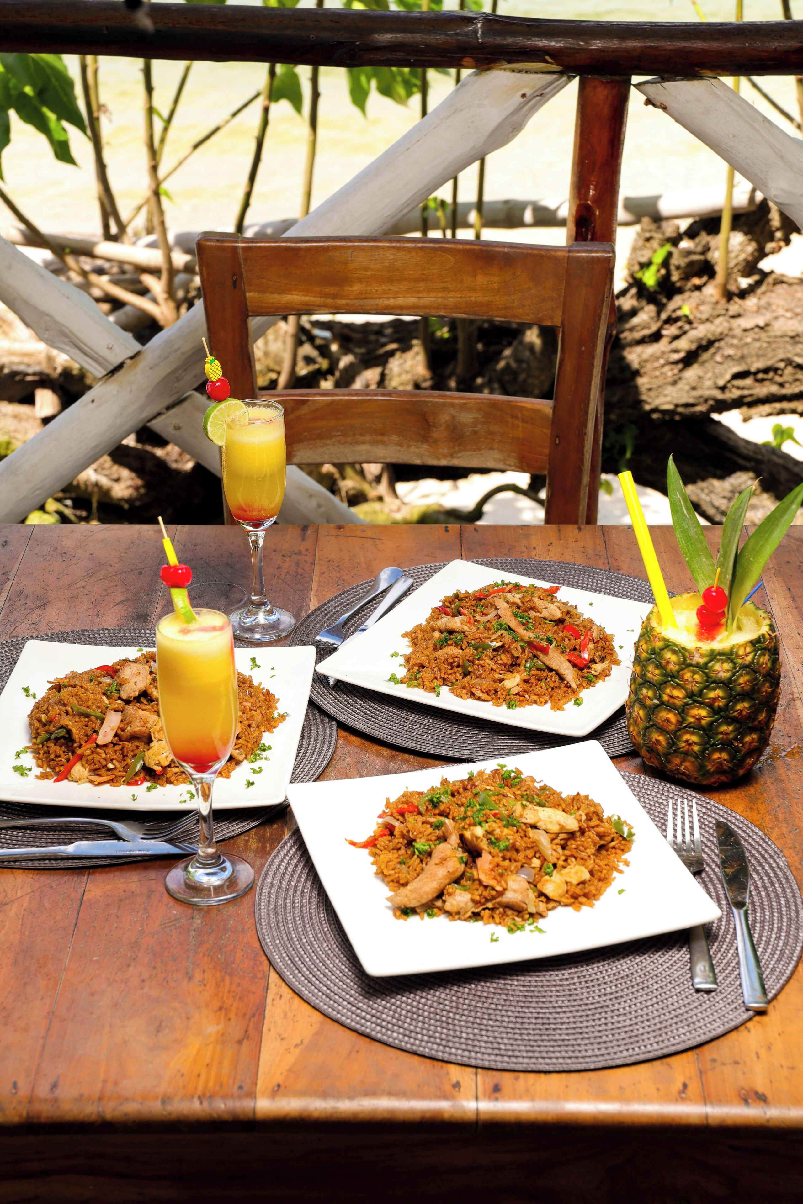 A wooden table with three square plates of fried rice, two glasses of a yellow beverage, and a pineapple with a decorative top.