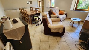A living room with a sofa, a dining table with chairs, a small table with a chair, and a window with a view of greenery. - Chez la Lise - TANCON (TANCON)