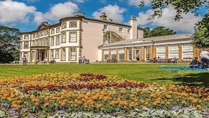 A large, historic building with a symmetrical facade, a central entrance, and a well-maintained lawn with colorful flower beds.