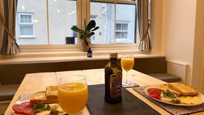 A breakfast setup with toast, tomatoes, cheese, and a bottle of juice on a table by a window with curtains.