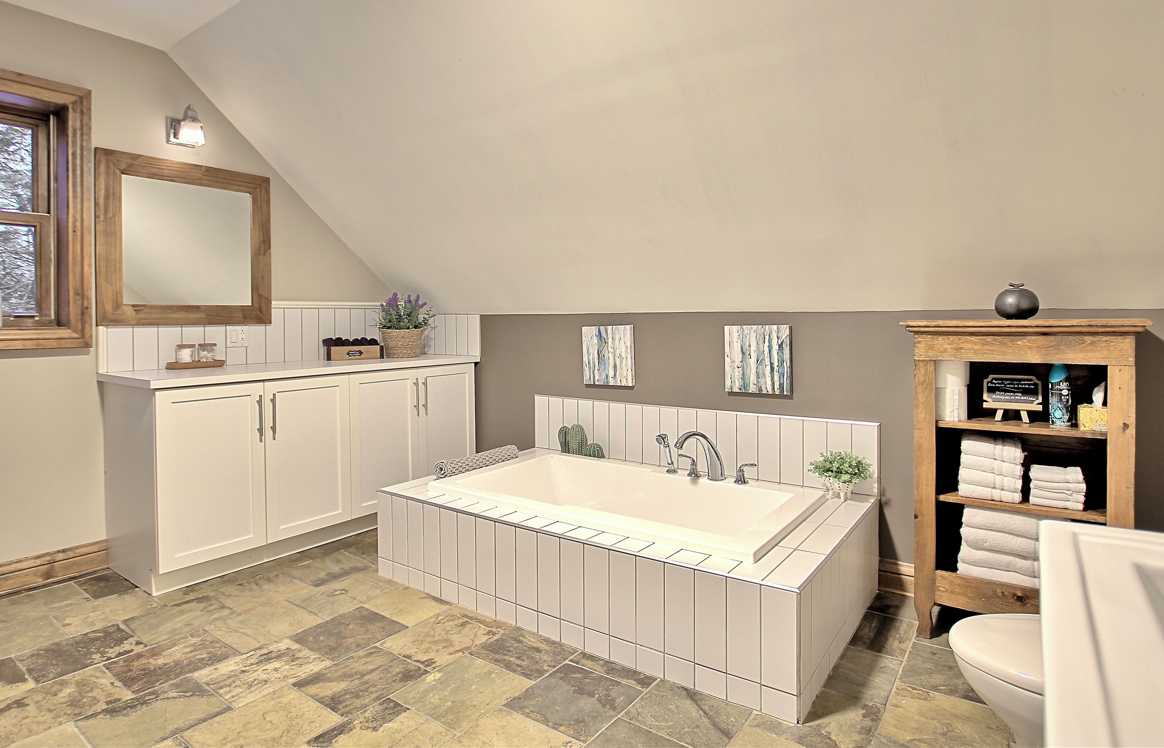 Un baño con una bañera grande, muebles de color blanco, un estante de madera con toallas y un espejo con marco de madera.