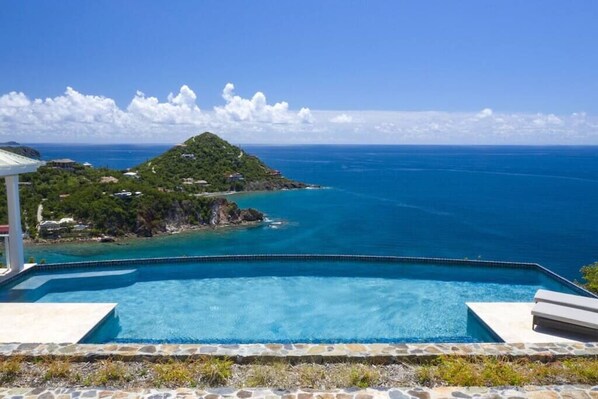 A clear view of a swimming pool with a stone border, overlooking a turquoise sea and a small island with lush greenery.
