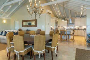 A spacious dining area with a large round table and wicker chairs, a chandelier, and a kitchen in the background. - Maria Bluff Estate | 7-Bedroom Villa | Generator (Cruz Bay)