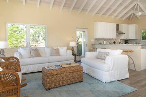 A living room with a sofa, a coffee table, and a dining area with chairs. There is a kitchen with cabinets and a window. - Maria Bluff Estate | 7-Bedroom Villa | Generator (Cruz Bay)