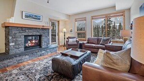 A cozy living room with a stone fireplace, leather sofas, and large windows offering mountain views. - Picturesque Views | Creekside Walking Paths to Town (Canmore)