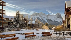 A snow-covered outdoor area with wooden benches and a stone pathway, surrounded by buildings and mountains in the background. - Picturesque Views | Creekside Walking Paths to Town (Canmore)