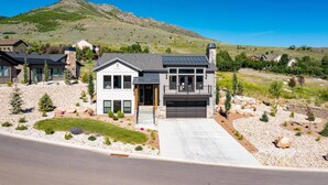 A modern two-story house with a garage, surrounded by a landscaped garden with small trees and shrubs, set against a backdrop of a hill and clear blue sky. - Private Powder Mtn B2 | Desk| Hot Tub| Ski Retreat (Eden)