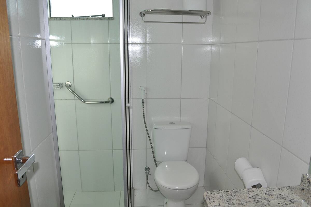 Um banheiro pequeno e limpo, com azulejos brancos, vaso sanitário, chuveiro com porta de vidro e porta-toalhas.