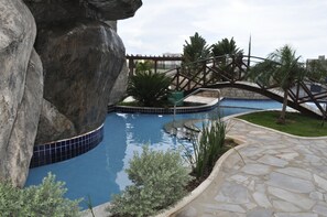 A swimming pool with a rock formation, a bridge, and a sign that reads "CINNA ADULTO". - Golden Dolphin Supreme - Achei Férias (Caldas Novas)