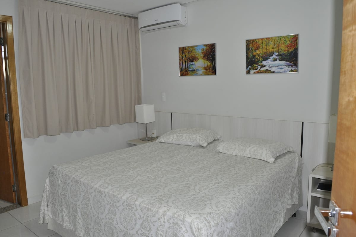 Uma cama bem arrumada com colcha estampada, uma mesa de cabeceira com abajur e duas pinturas na parede.
