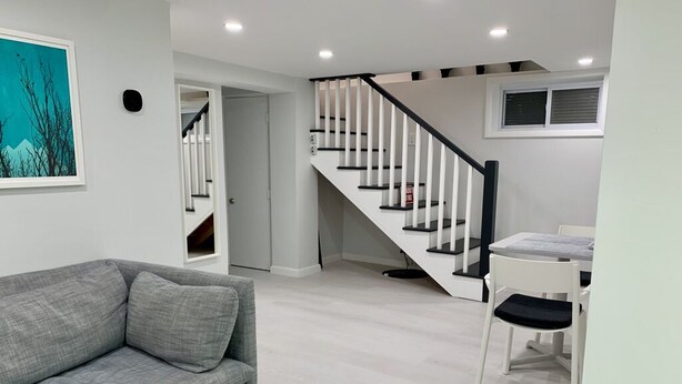 Een moderne woonkamer met een grijze bank, een trap met zwarre leuningen, en een eethoek met een witte tafel en stoelen.