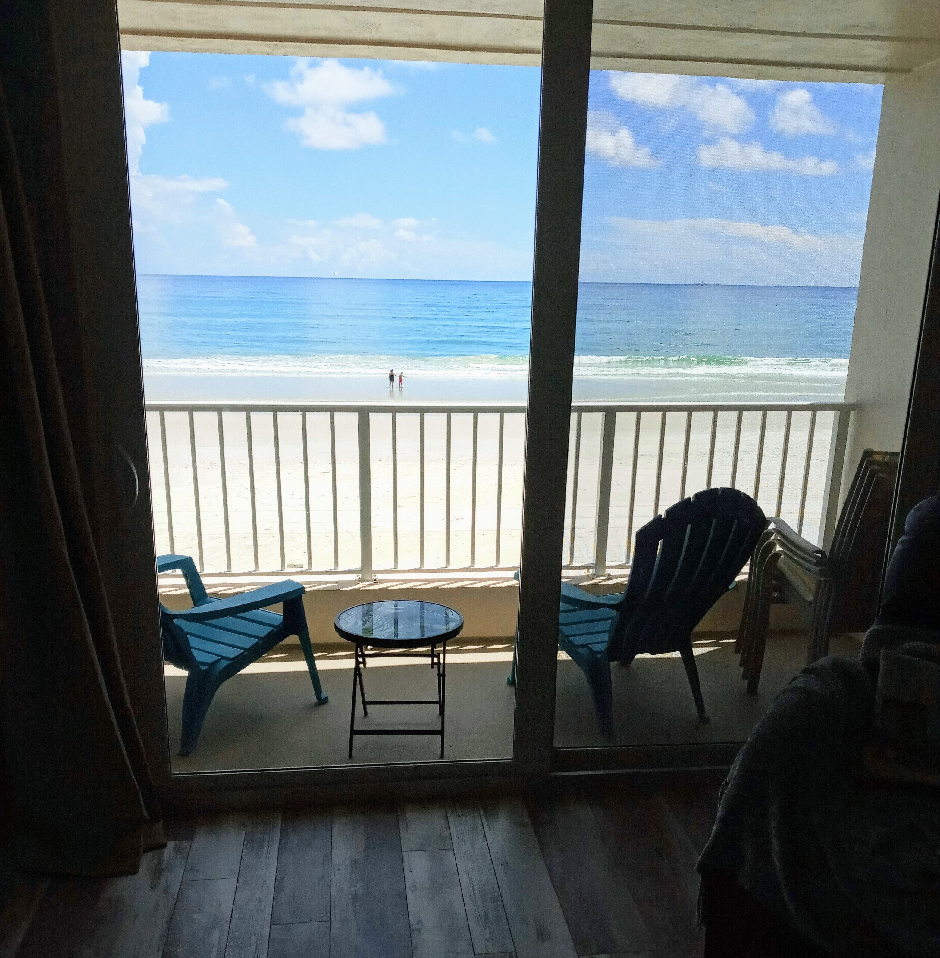 Un balcón con vista a la playa, dos sillas azules y una mesa redonda pequeña.