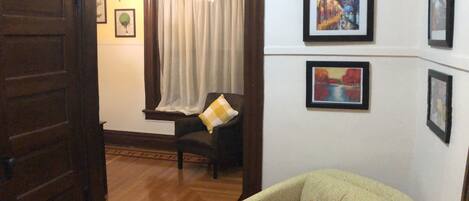 A room with a wooden door, a patterned rug, a chair with a cushion, and framed pictures on the wall.