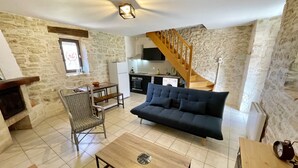 A cozy living room with a sofa, a wooden coffee table, and a stone wall. A kitchen with a wooden staircase is visible in the background. - Cottage near Barjac (Saint-Privat-de-Champclos)