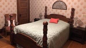 A bedroom with a wooden four-poster bed, a red pillow, a nightstand with a lamp, and a chair with a cushion.