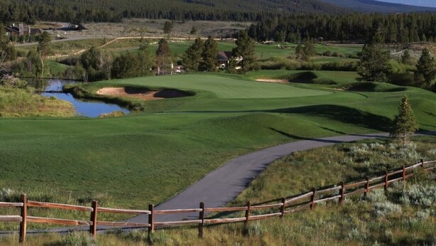 En golfbane med et træhegn, en sø og bjerge i baggrunden.