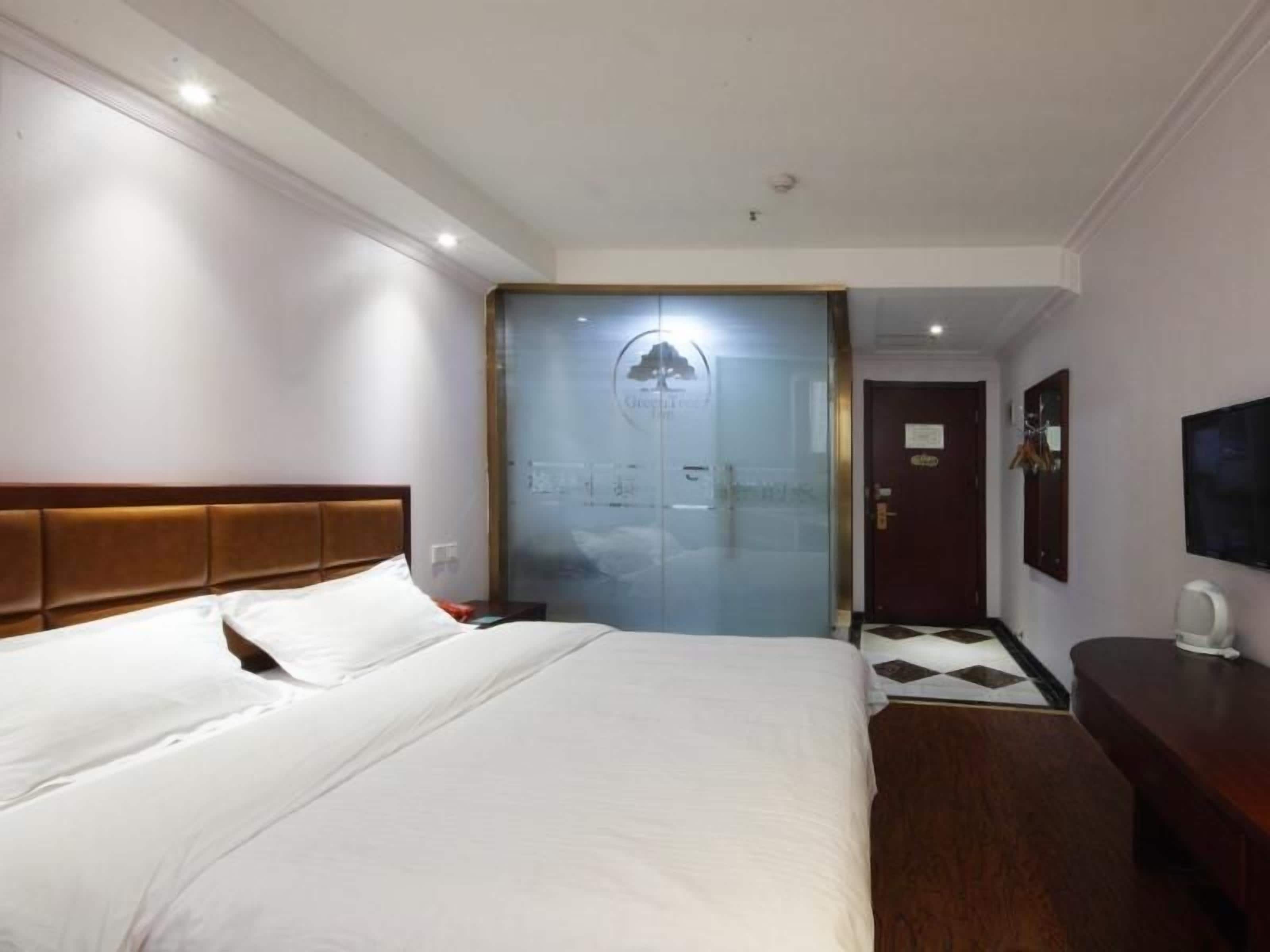 A hotel room with a large bed, a wooden headboard, a desk with a chair, a television, and a glass door leading to a hallway.