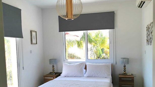 A bedroom with a large bed, a hanging light fixture, a window with a view of greenery, and a small bedside table with a lamp. - Villa Alba, Luxury 4 BDRM Villa with Heated Pool (Protaras)