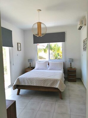 A bedroom with a large bed, a hanging light fixture, a window with a view of greenery, and a small bedside table with a lamp. - Villa Alba, Luxury 4 BDRM Villa with Heated Pool (Protaras)