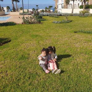 Deux enfants jouent sur une pelouse, avec une piscine et des palmiers en arrière-plan.