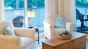 Een ruime woonkamer met een plafondventilator, twee witte fauteuils, een houten salontafel en uitzicht op een balkon met groen.