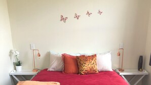 Een net opgemaakt bed met een rode sprei, witte kussens en een decoratief oranje kussen. Er staan twee nachtkastjes met lampen, een kleine potplant en wanddecoratie met vlinders.