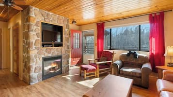 A cozy living room with a stone fireplace, wooden ceiling, and a large window with red curtains.