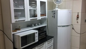 A kitchen with a microwave, oven, and refrigerator. There are cabinets above the counter and a fan on the wall. - Apartamento de um Quarto (RJ)