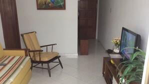 A living room with a sofa, a chair, a TV, and a small table with a vase of flowers. - Apartamento de um Quarto (RJ)