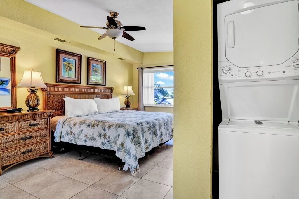 Quarto de hotel com cama, ventoinha no teto, janela com vista, cómoda e máquina de lavar roupa.