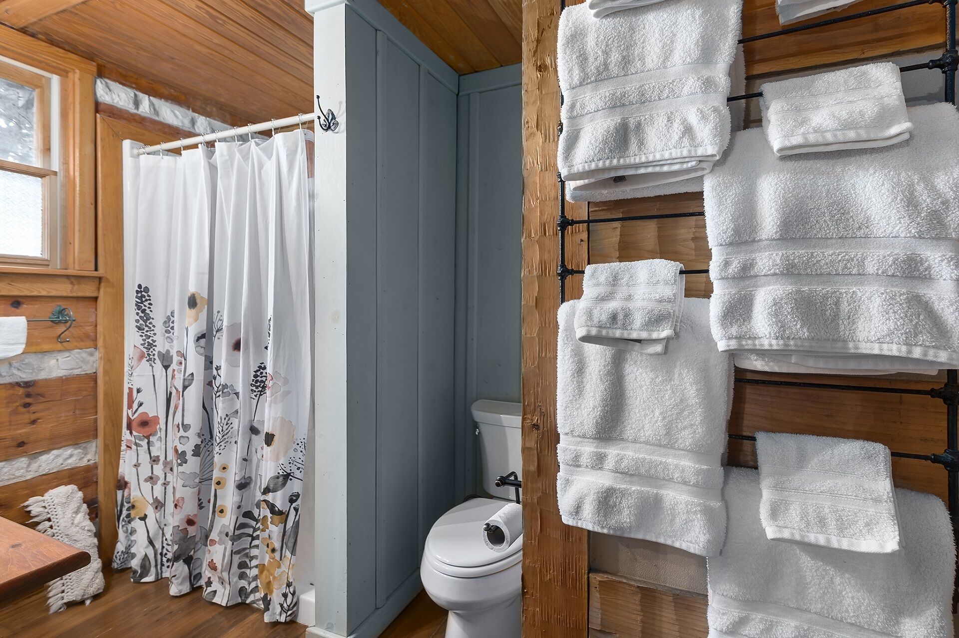 Ein Badezimmer mit Holzvertäfelung, einer Duschvorhang mit floralem Muster und einem Handtüchhalter mit mehreren weißen Handtüchern.